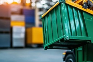 large green dumpster is being lifted by machinery within a warehouse environment