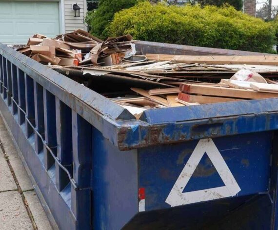blue dumpster full of wood and other debris in the driveway in front of a house in the suburbs