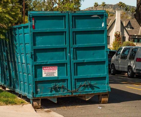 large teal dumpster bin parked on a residential street with cars and houses in the background