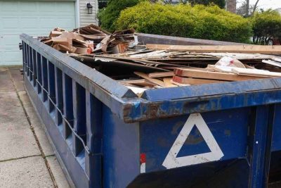 blue dumpster full of wood and other debris in the driveway in front of a house in the suburbs