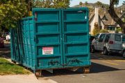 large teal dumpster bin parked on a residential street with cars and houses in the background
