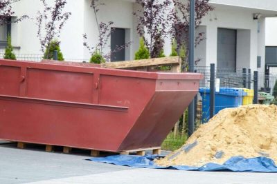 red construction dumpster next to a pile of sand on a tarp in front of modern residential buildings